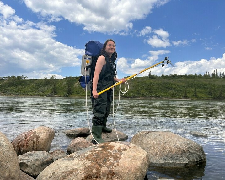 Young woman standing on a rock beside a waterbody, wearing a qPCR sampling backpack.