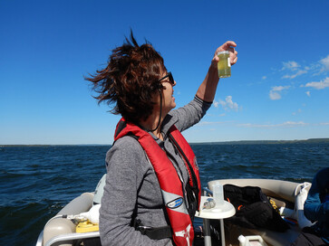 Woman in lifejacket holding vial on boat with lake in the background.