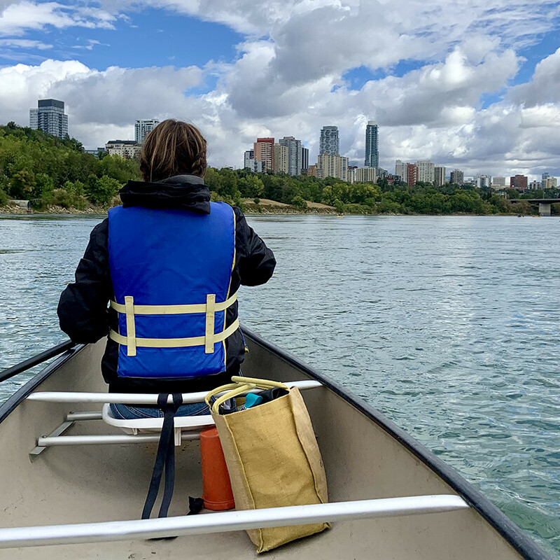 Woman paddling in canoe with clear water and blue skies with Edmonton's skyline in the background.